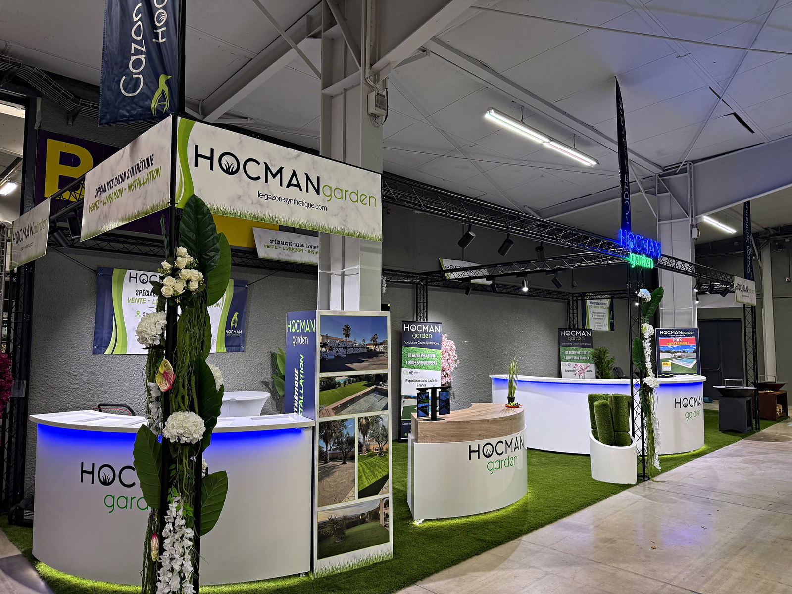 Stand Hocman Garden sur un salon : comptoirs blancs éclairés, sol en gazon synthétique et rouleaux exposés