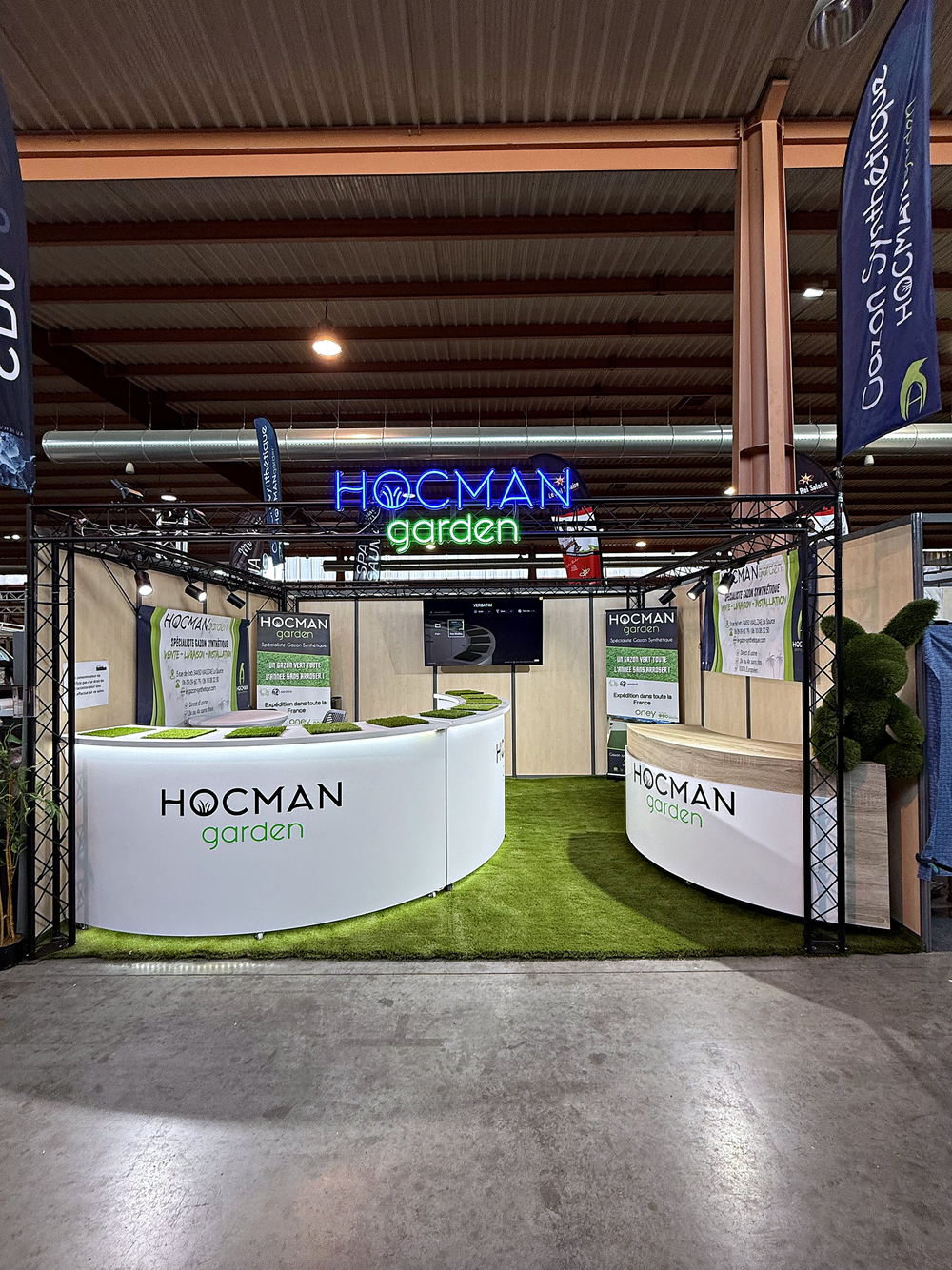 Stand Hocman Garden vu de l'allée : enseigne néon et échantillons de gazon synthétique sur le comptoir
